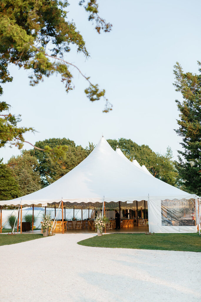 An expansive wedding tent set up in an outdoor area with a tree-lined pathway leading to it, under a clear sky at dusk.
