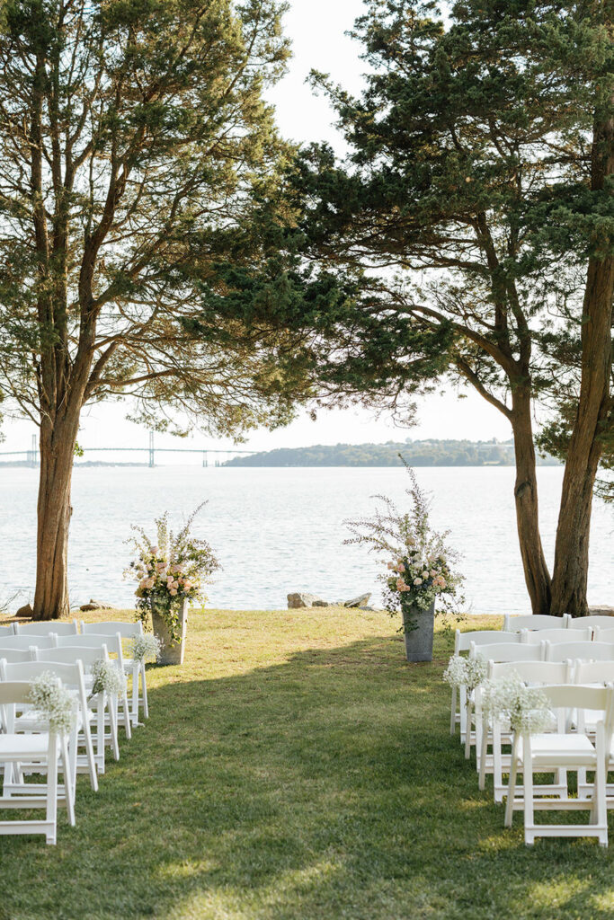 Rows of white chairs set up for a wedding ceremony by the waterfront, flanked by trees and floral arrangements, creating a serene outdoor venue.