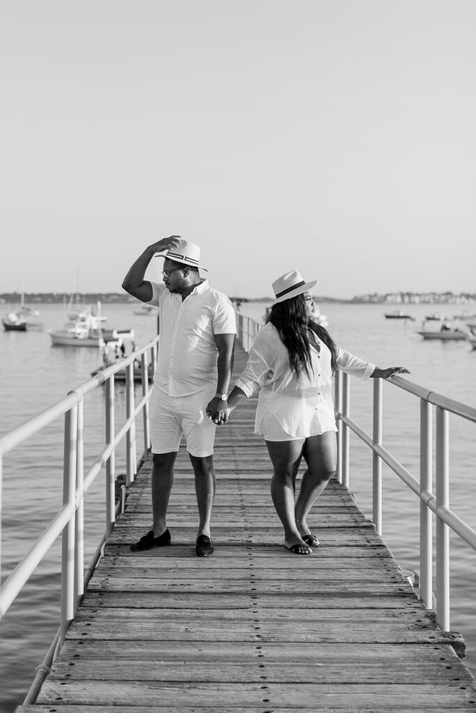 A stylish couple in white outfits holding hands on a wooden pier, with boats in the background, evoking a serene maritime atmosphere