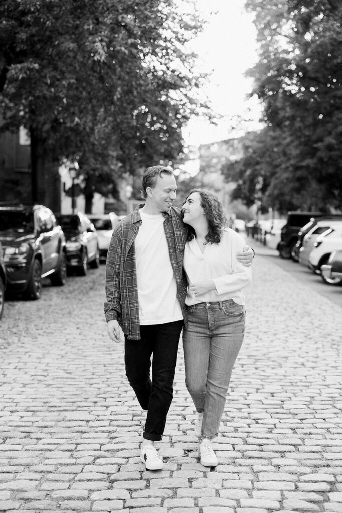 A loving couple walking arm-in-arm down a picturesque cobblestone street, exuding warmth and closeness in their casual attire.
