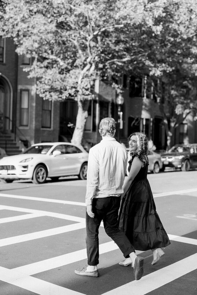 A couple walking away, crossing a zebra crossing on a sunny day, with one turning back and smiling, amidst a street lined with cars and trees