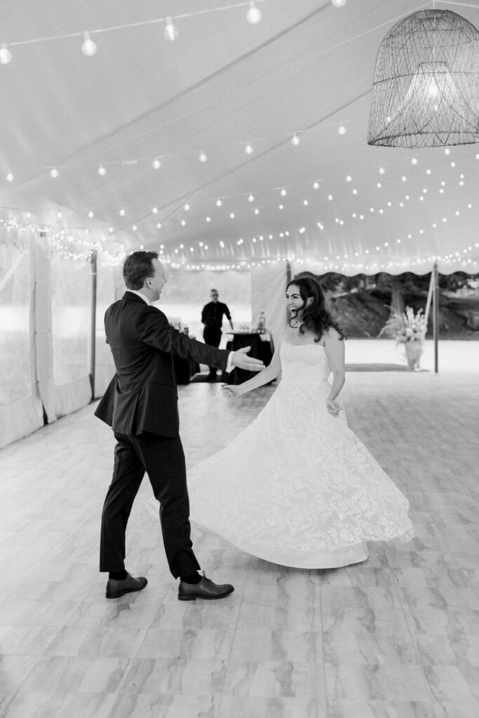 A black and white photograph capturing a moment of joy as the bride twirls in her lace gown while dancing with the groom under a canopy of string lights at the reception.