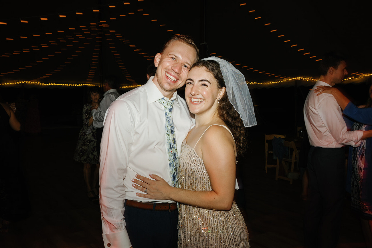 A newlywed couple shares a cheerful moment on the dance floor; the bride in a sparkling golden dress and a veil, and the groom in a white shirt and patterned tie, both smiling widely under festive string lights.