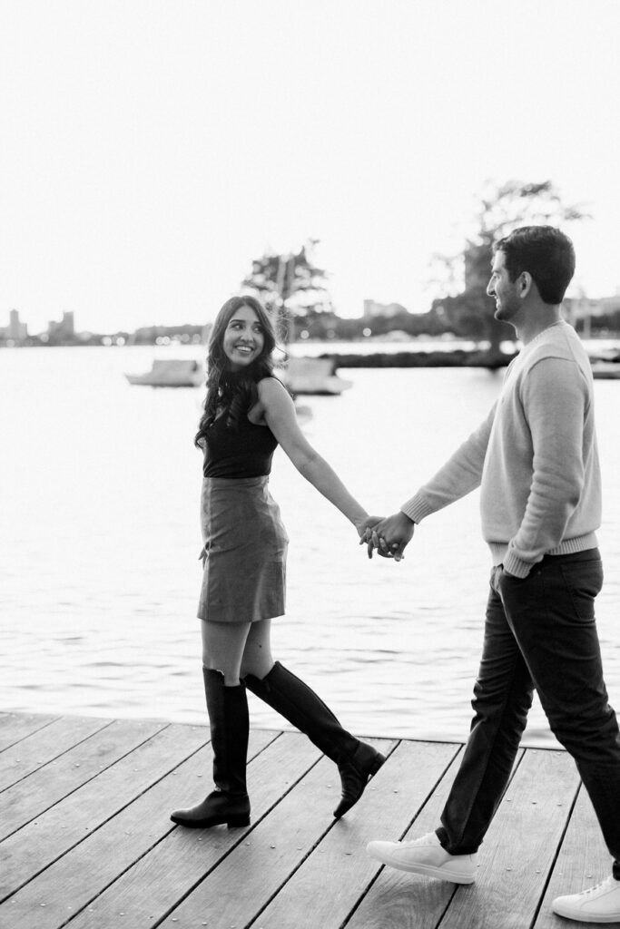 A playful black and white photograph captures a couple walking hand in hand along the Boston Esplanade, with boats and the city skyline blurred in the background.