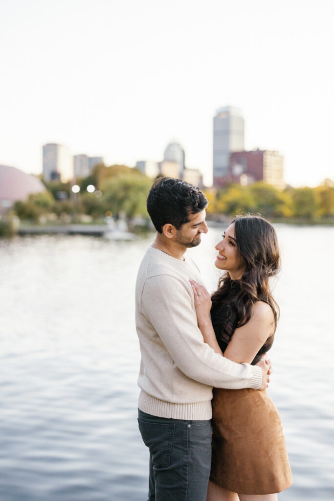 A couple embraces lovingly on the Boston Esplanade, with the city skyline and water behind them, creating a serene and romantic atmosphere