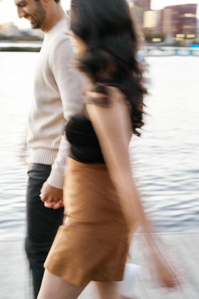 A dynamic image of a couple in motion, the woman's hair and skirt fluttering as they walk briskly, their hands clasped, along the Boston Esplanade.