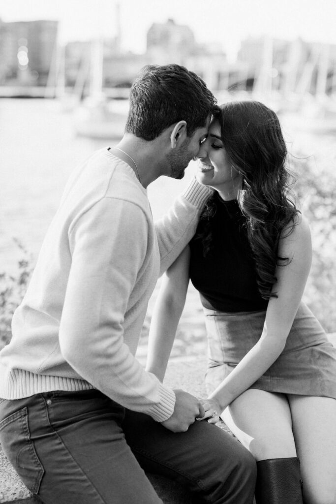 An intimate black and white portrait of a couple sitting close together by the waterfront, their faces touching tenderly, exuding love and closeness.