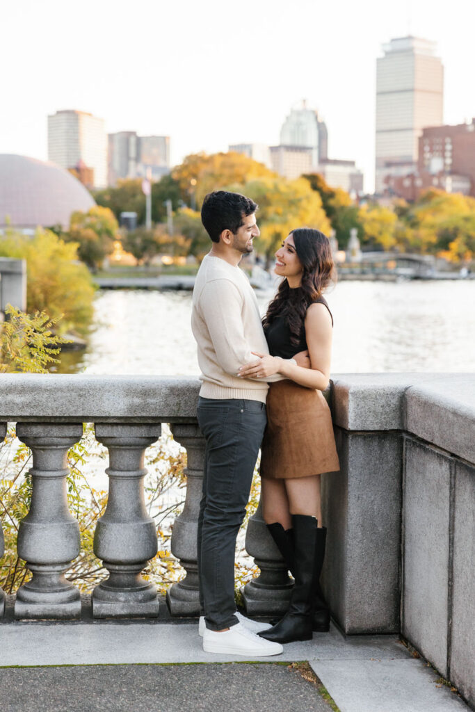 A couple leans lovingly against a stone balustrade overlooking the Charles River Esplanade, with the Boston skyline in the background, sharing a serene moment.