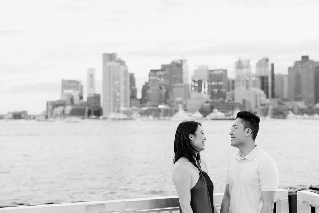 A couple holding hands against a fence overlooking the water and Boston city skyline.
