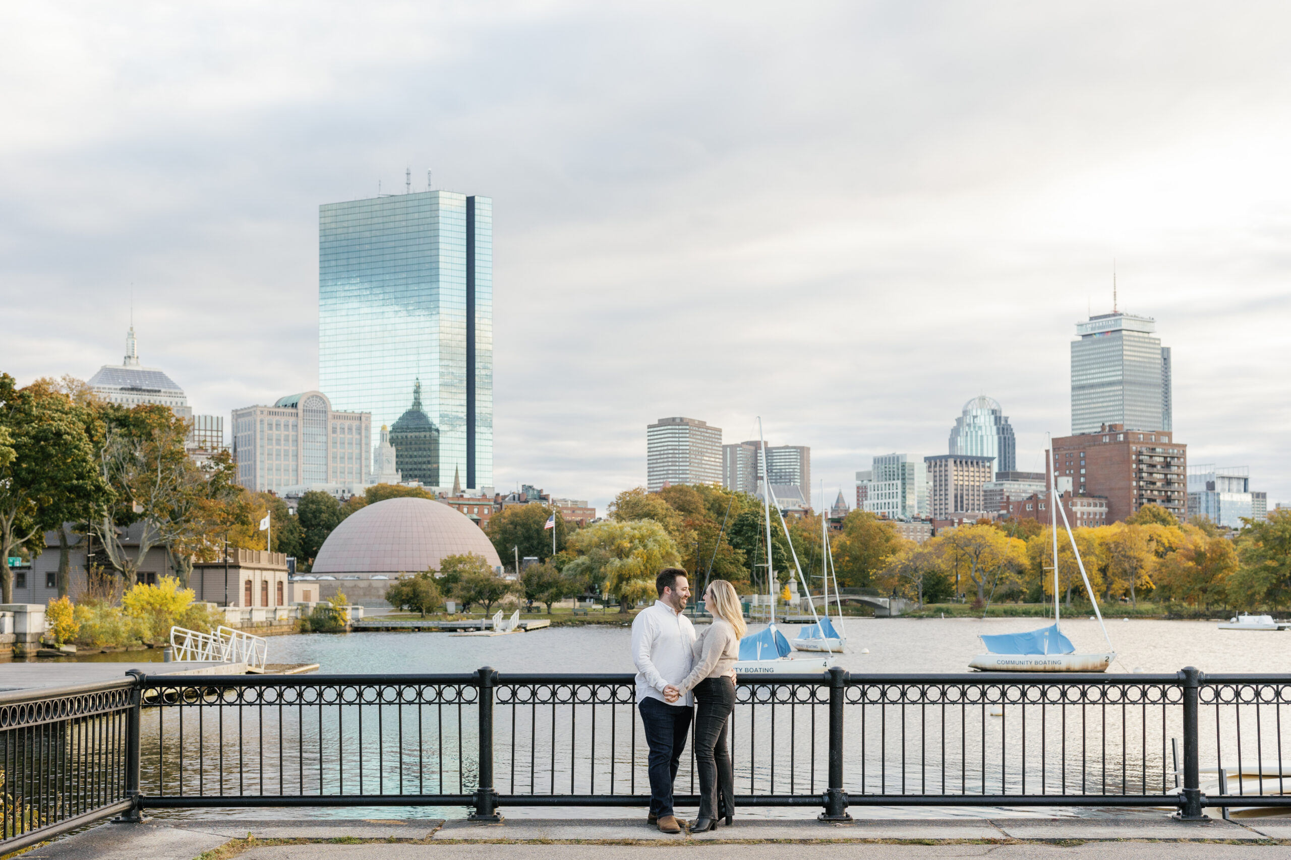 A romantic couple standing close, with a city riverfront and autumnal trees in the soft glow of the setting sun as their backdrop.