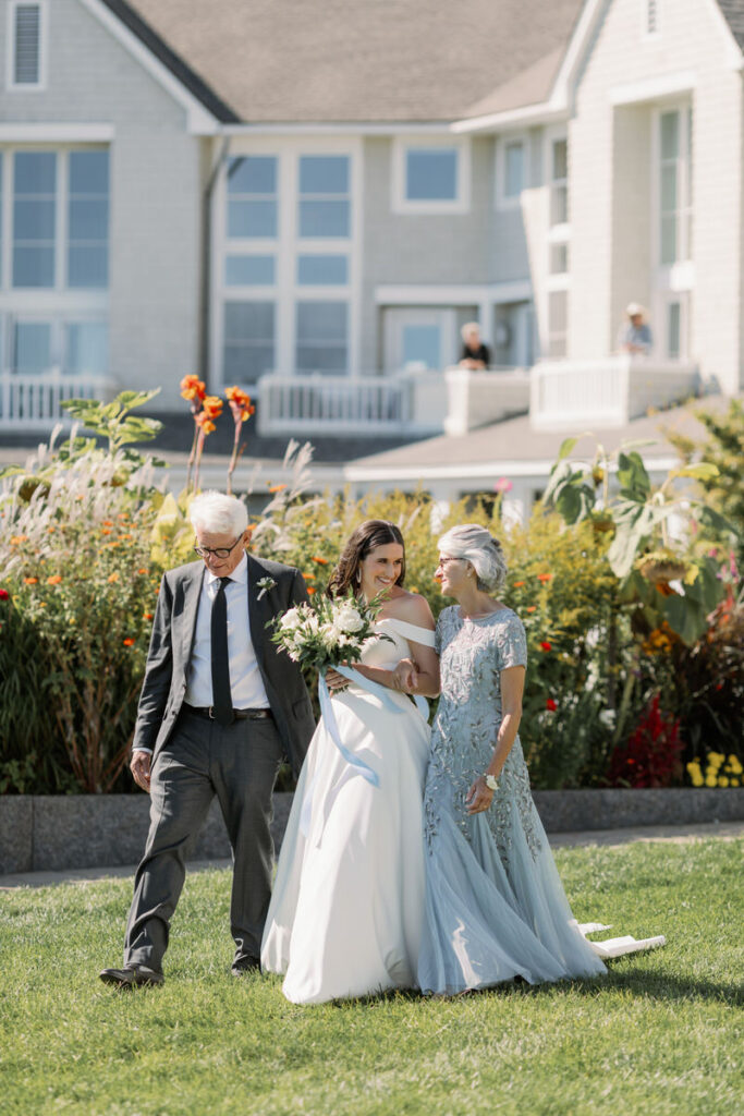 Bride with her parents, walking and smiling in a sunny outdoor wedding ceremony with a house in the background