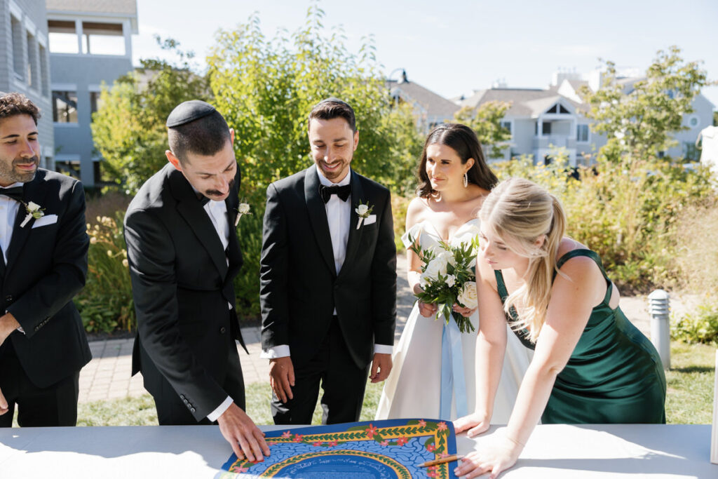 Wedding guests looking at a colorful Ketubah signing board during a ceremony, with a backdrop of lush greenery