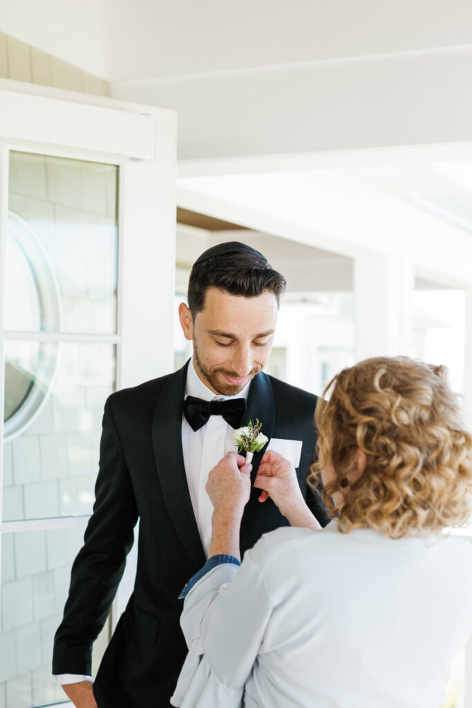 Groom in a black tuxedo having a white boutonniere pinned to his lapel by a woman in a light blue dress
