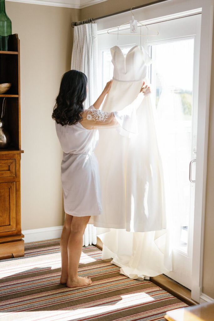 Bride in a white robe admiring her wedding dress hanging in front of a bright window