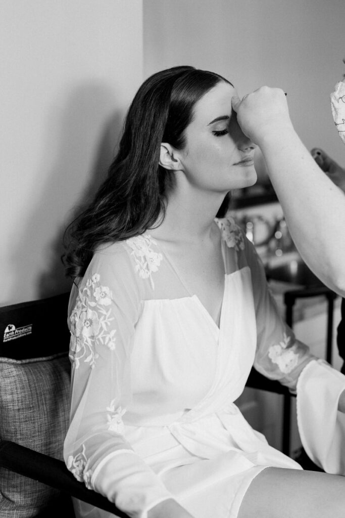 Bride in a white robe with floral embroidery getting makeup applied before the wedding