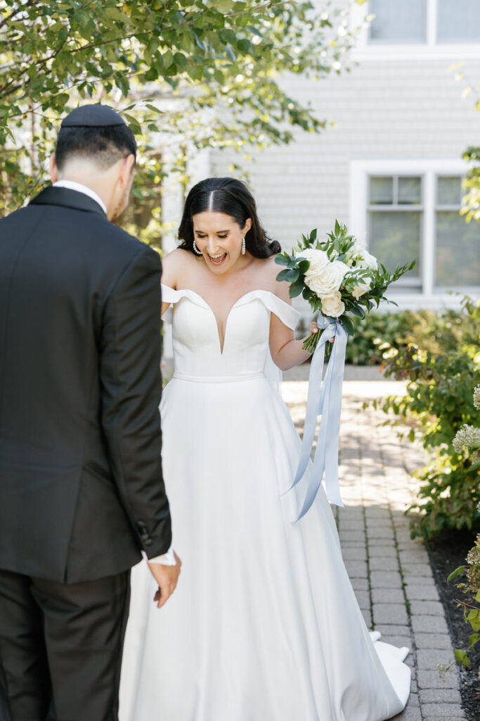 Excited bride in a white off-shoulder wedding dress, laughing as she looks at the groom outdoors