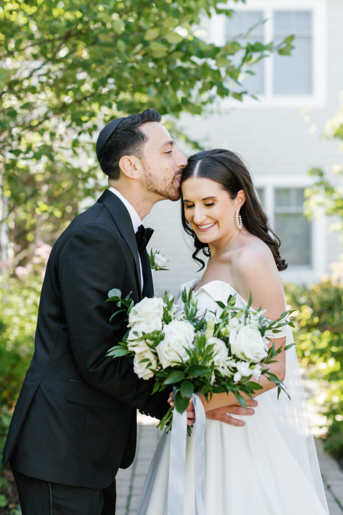 Groom in a black tuxedo and kippah kissing the head of the smiling bride holding a bouquet