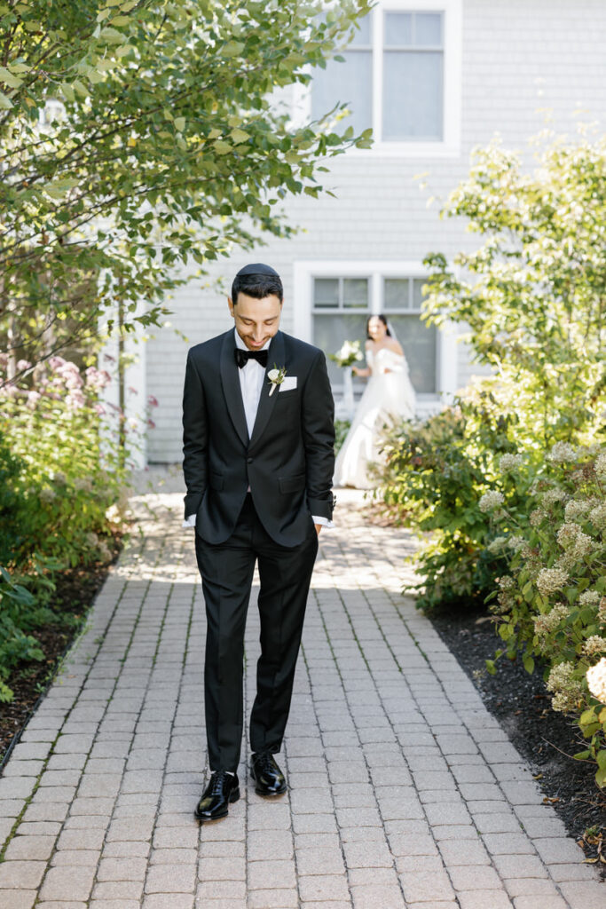 Groom in a black suit looking down a path with the bride approaching in the background