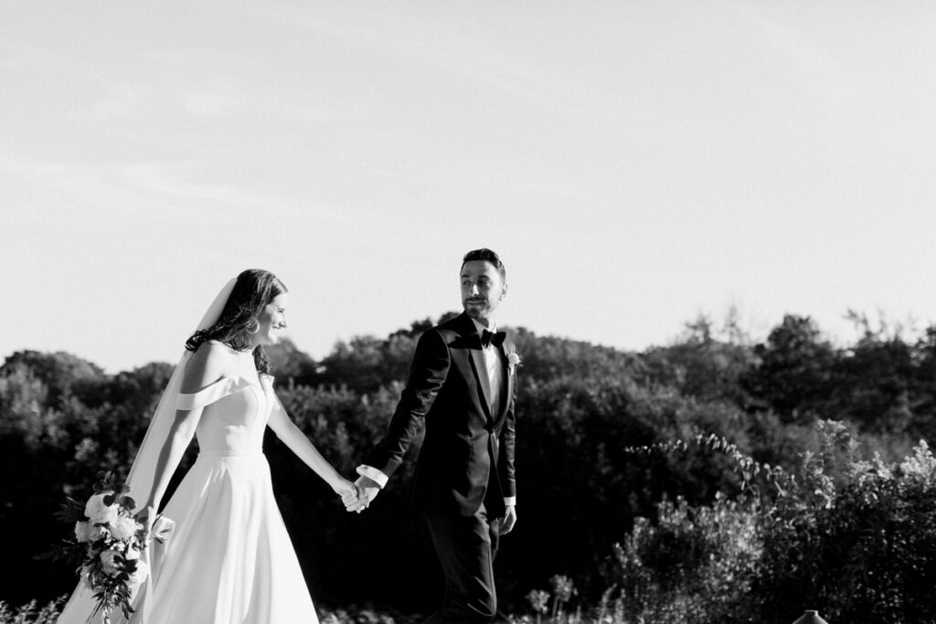 Black and white image of a bride and groom holding hands, walking through a field