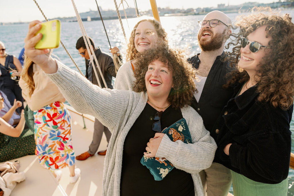 A group of 3 women and 1 man takie a selfie together on a boat while the wind blows their hair