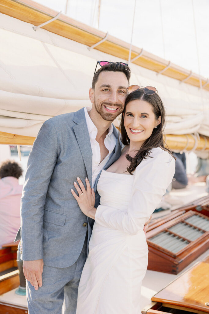 Happy couple smiling on a sailboat, the man in a gray suit and the woman in a white dress, both wearing sunglasses
