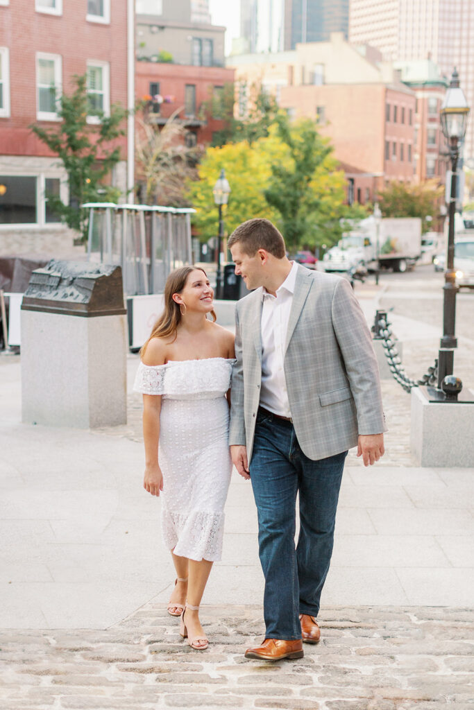 A couple looking at each other smiling as they walk along a sidewalk in a city. 