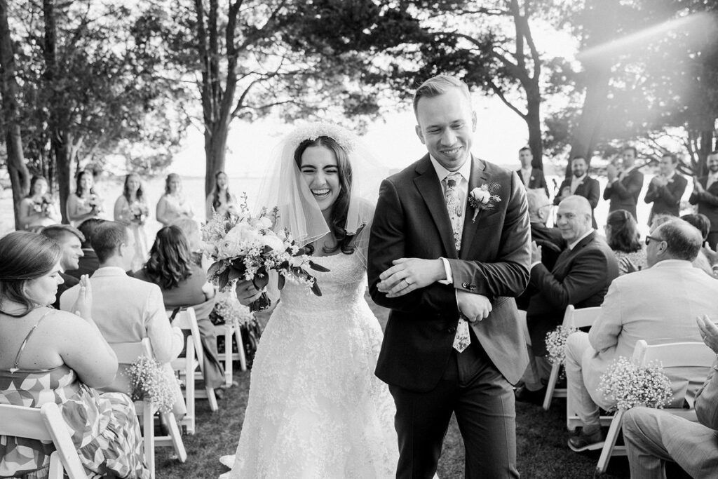 Black and white photo of a bride and groom walking hand in hand down the aisle, with wedding guests seated on either side and trees in the background