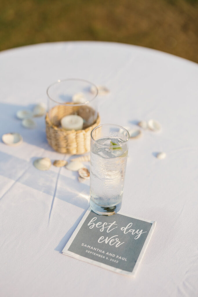 Close-up of a cocktail table centerpiece featuring a glass of water with lime, a wicker basket with white candles, and scattered seashells, with a 'best day ever' card including names and date