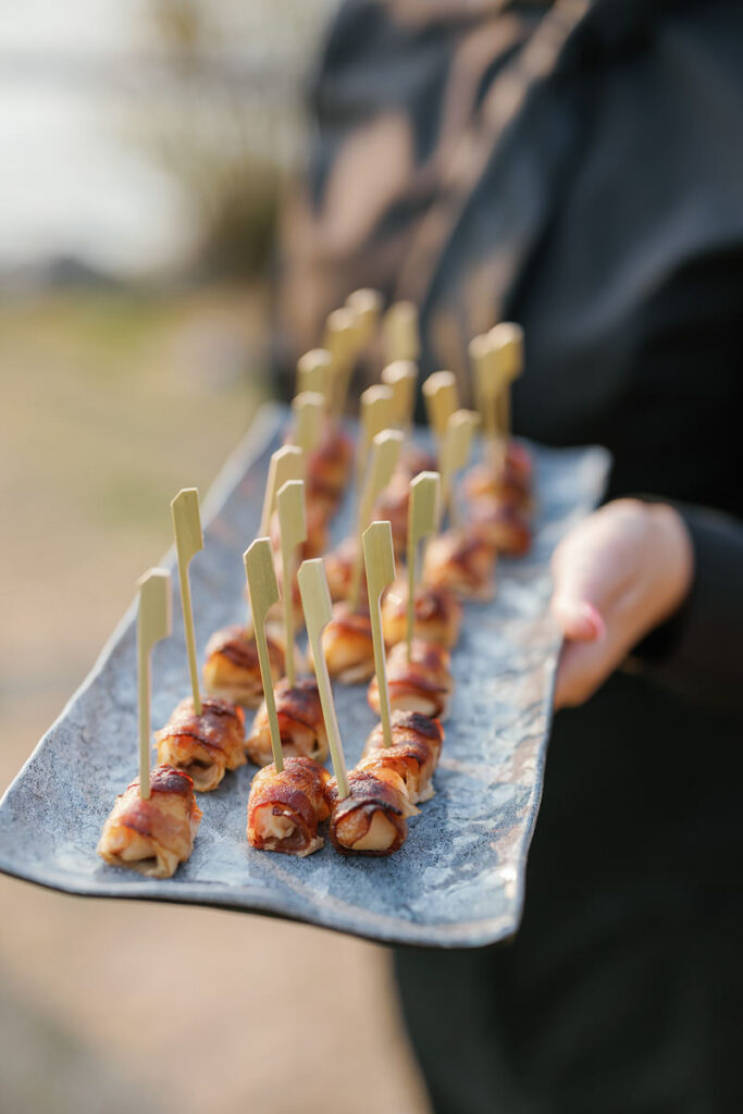 A caterer presenting a tray of bacon-wrapped dates, each skewered with a wooden toothpick, arranged neatly on a blue marbled platter