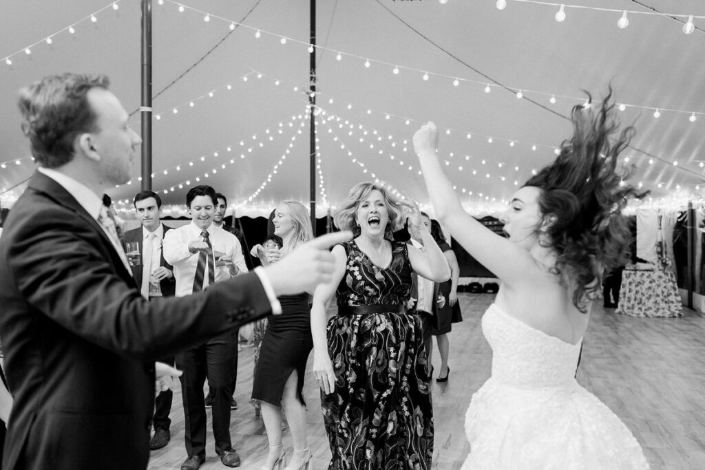 Black and white photo showing a dance floor at a wedding reception with guests dancing, a bride spinning with her hair flying, under a canopy of string lights