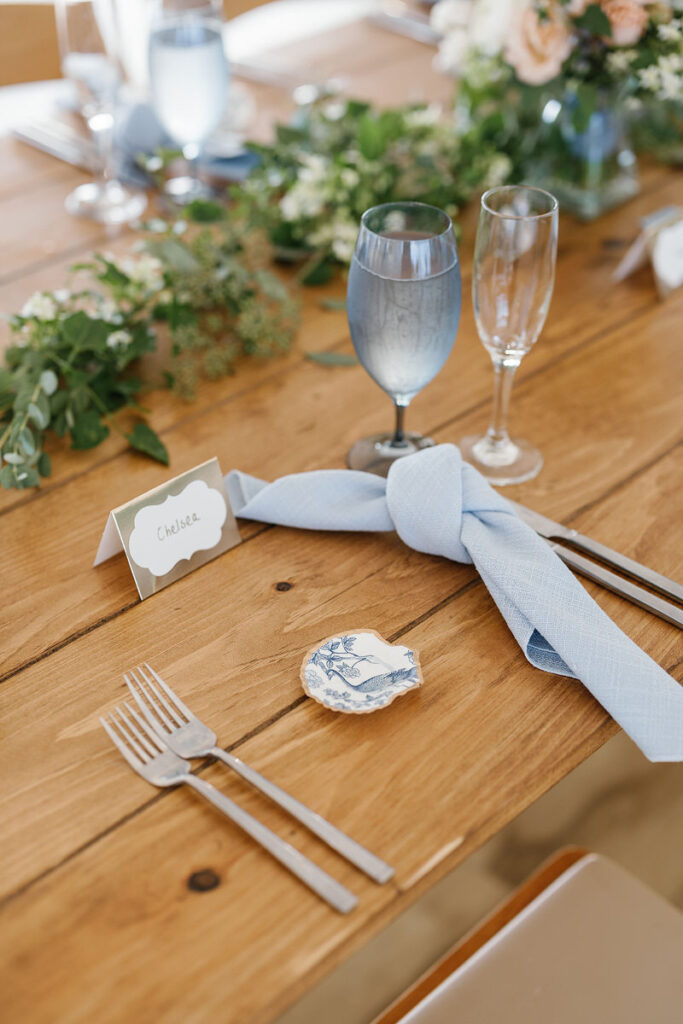 A detailed view of a wedding reception table with a light blue napkin, silverware, a small floral decor piece, and a name card reading 'Chelsea'