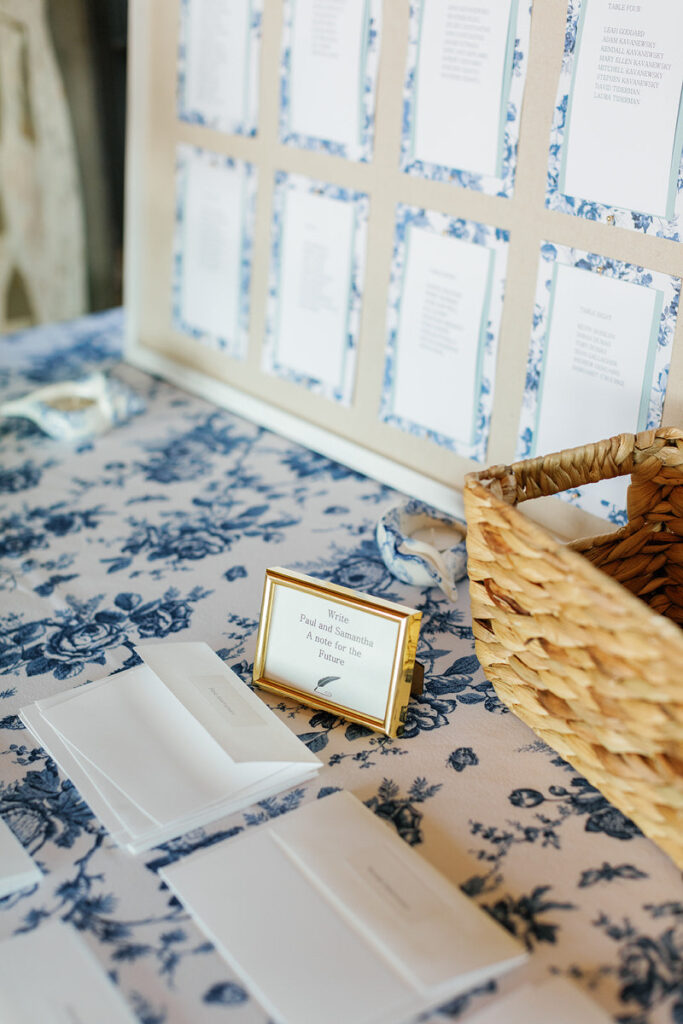 A framed wedding seating chart with blue floral borders displayed on a table with a matching blue floral tablecloth, accompanied by cards and a woven basket