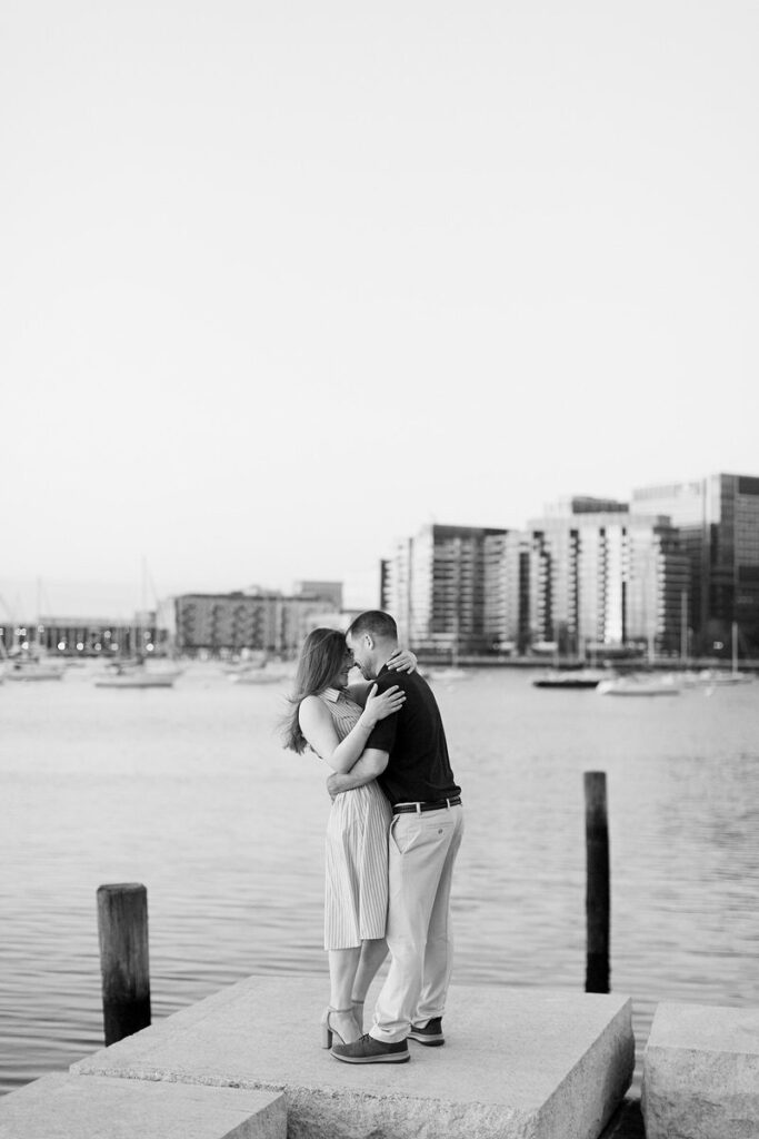 A couple embracing on a waterfront dock in a black and white photo, with boats and city buildings in the background