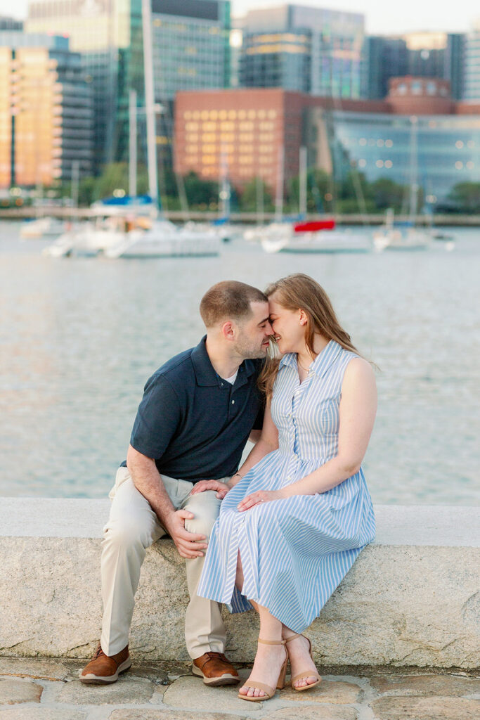 Two people sitting closely on a waterfront ledge, about to kiss with a cityscape and moored boats behind them