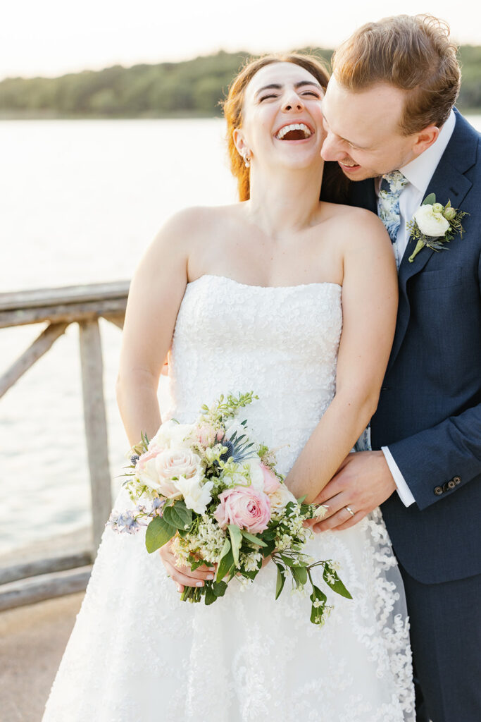 A beaming bride in a strapless white gown laughs joyously while the groom, in a blue suit, embraces her from behind by the waterfront, both enjoying an intimate moment.