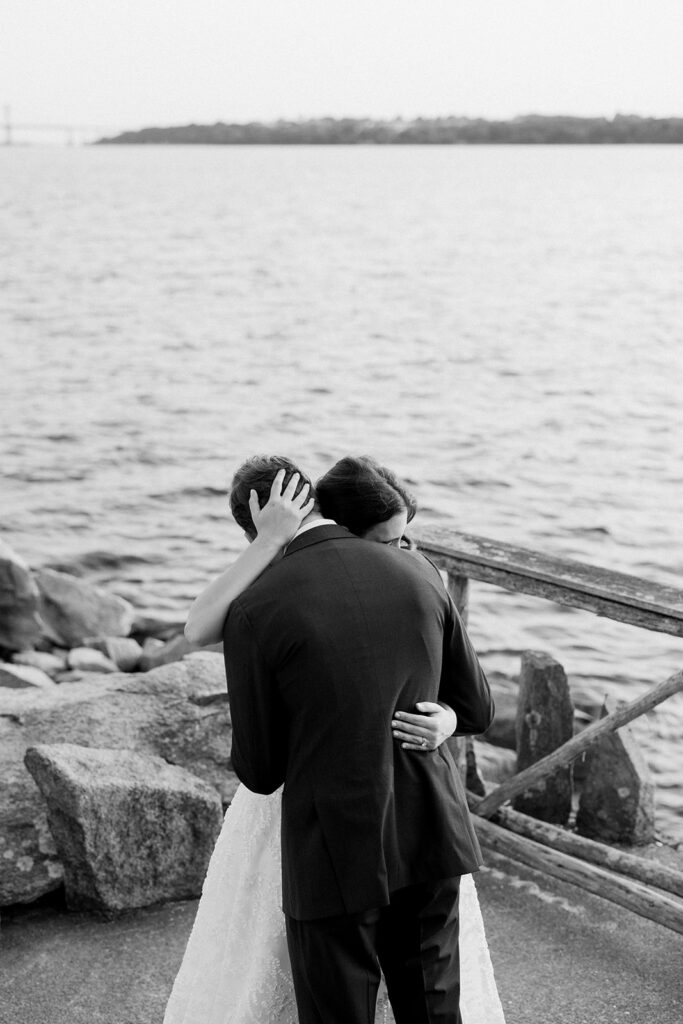 A black and white image of a bride wrapping her arms around the groom and embracing in a hug by the ocean.