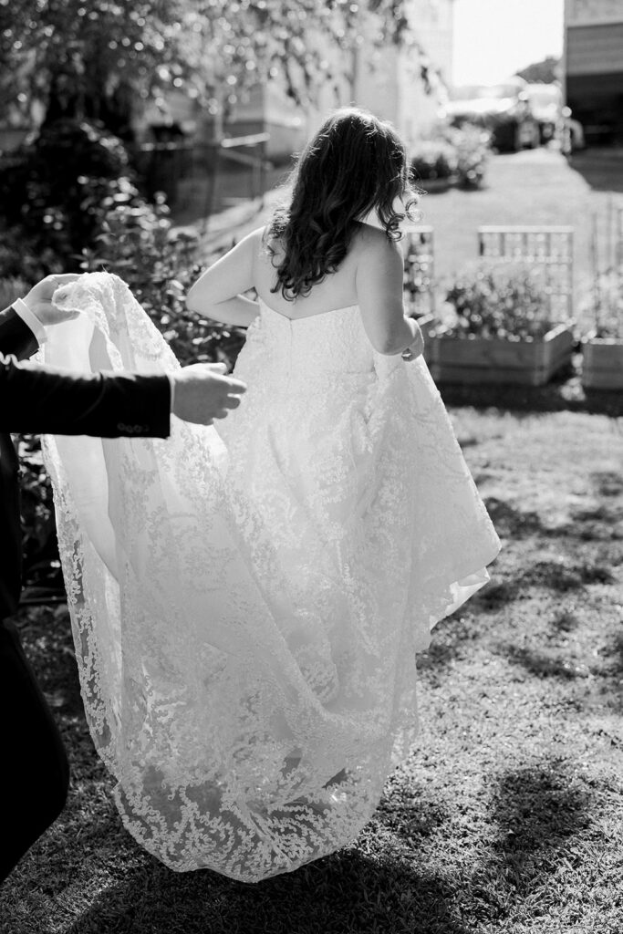 A black and white photo of a bride walking away holding up her dress with the help of the groom.