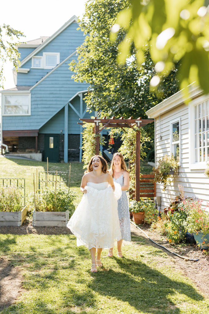 A smiling bride approaching through garden with a bridesmaid holding up the back of her dress.