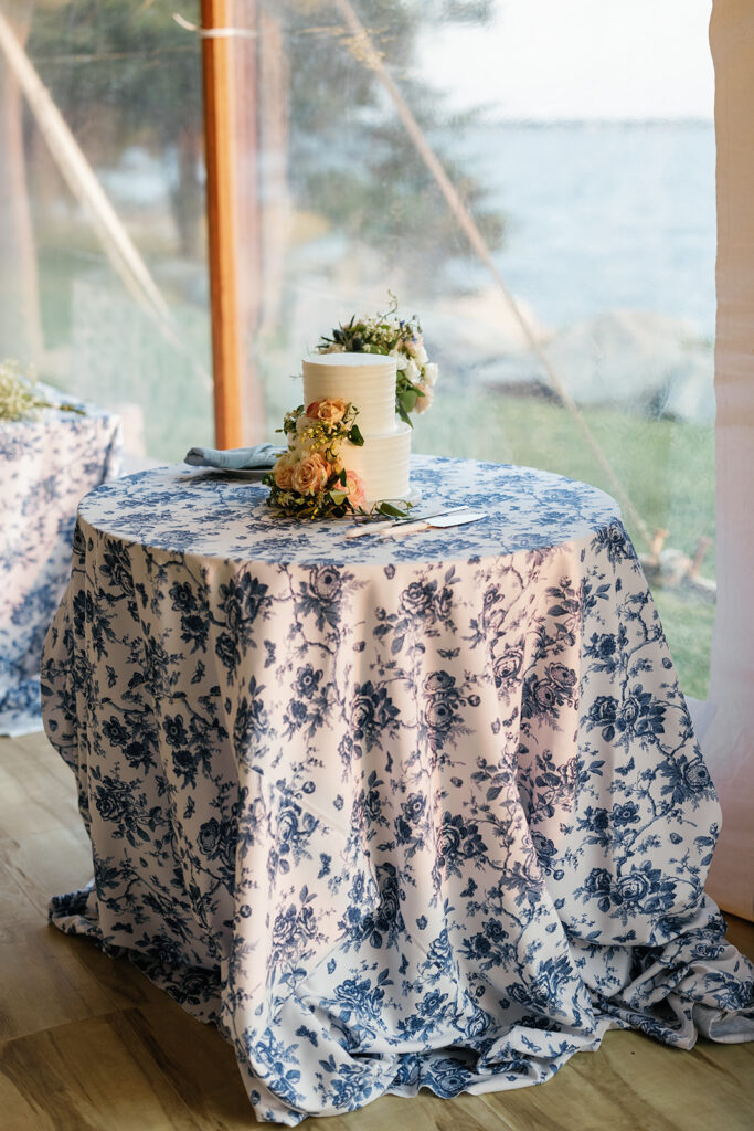 Small two tiered white cake decorated with flowers on a blue floral table cloth.
