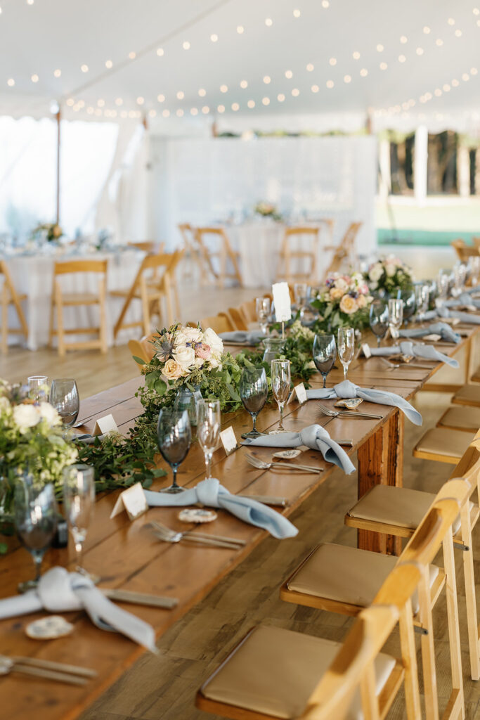 Long wooden farm table set up under a white tent and the table is adorned with blue tied napkins, blue glasses, and floral arrangements.