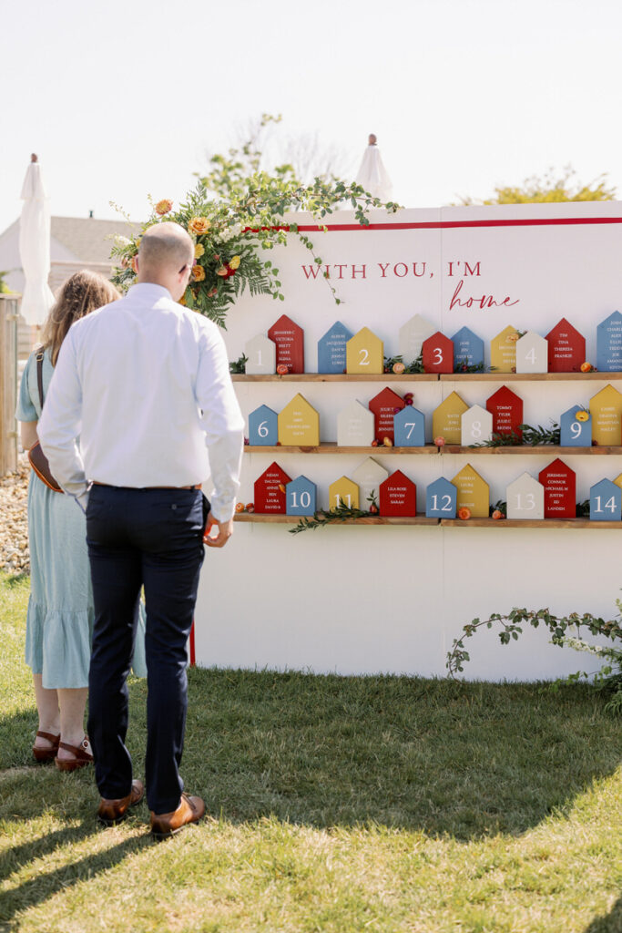 Two wedding guests looking at a wall with table placements on it. 
