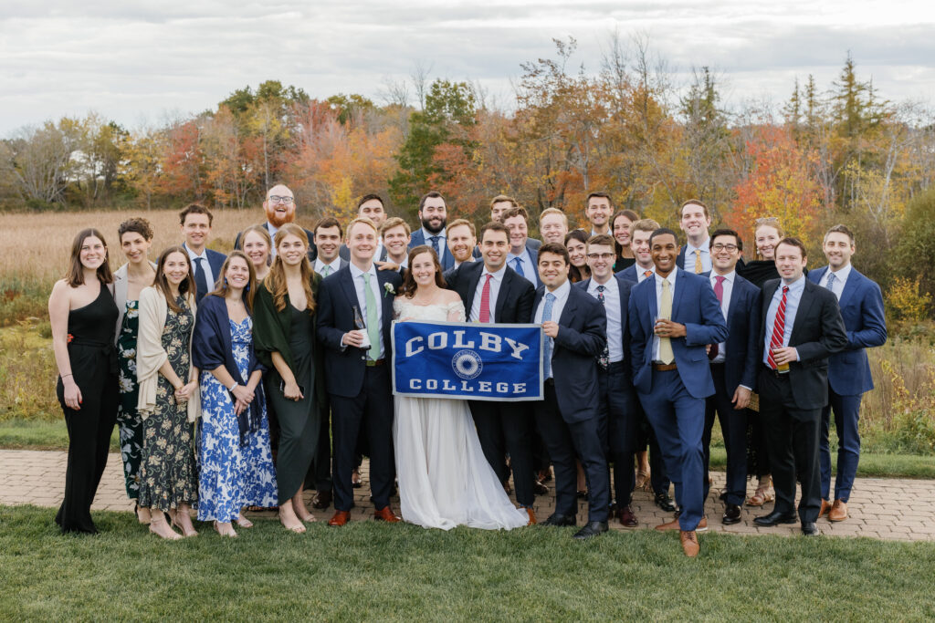 The bride and groom hold a "Colby College" flag while surrounded by wedding guests