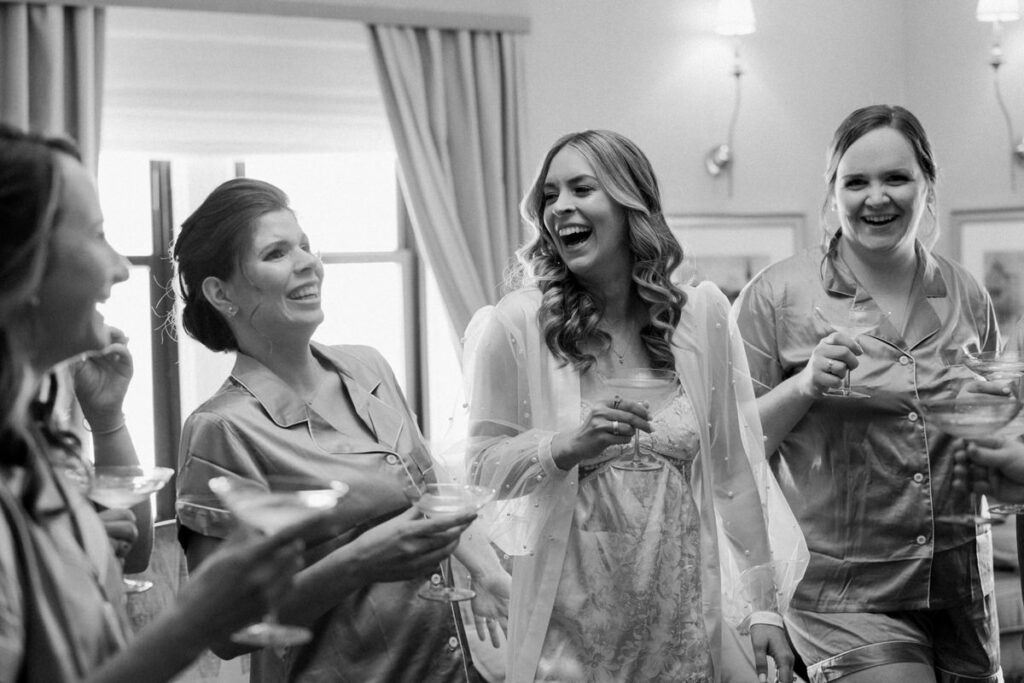 A bride and bridesmaids standing together holding glasses of champagne and laughing.