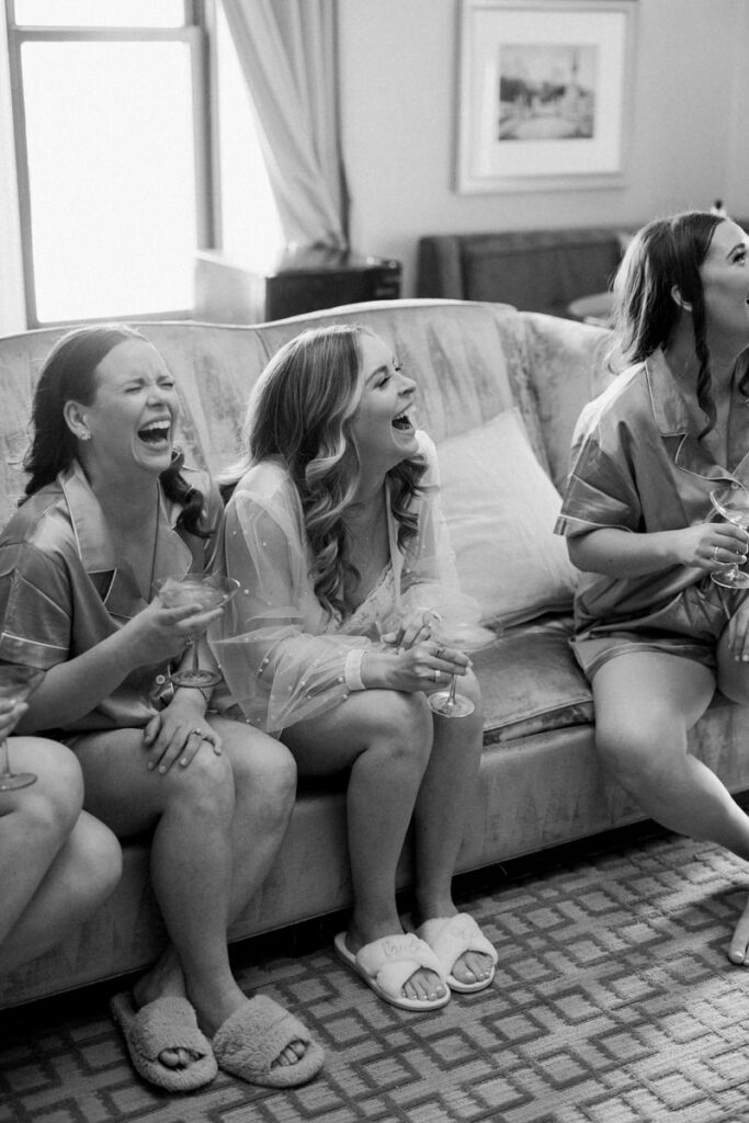 A bride and bridesmaids sit on a couch laughing 