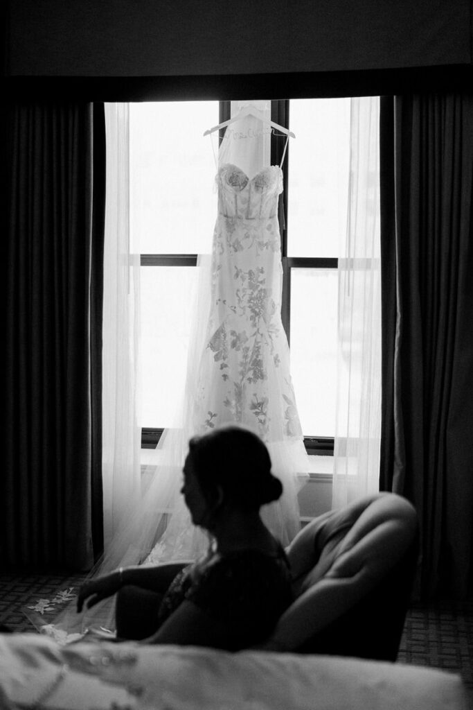 Silhouette of a person sitting on a couch with a wedding dress hanging in a window behind. 