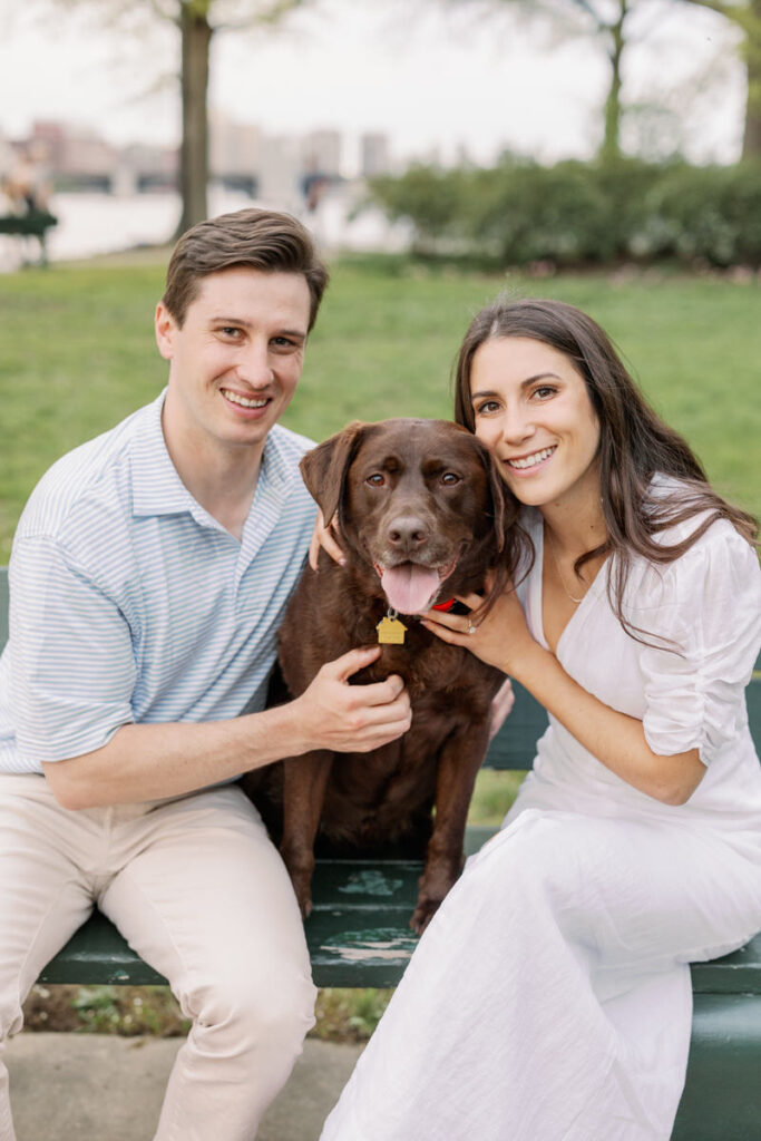 Two people sitting on a bench smiling with a dog in between them. 