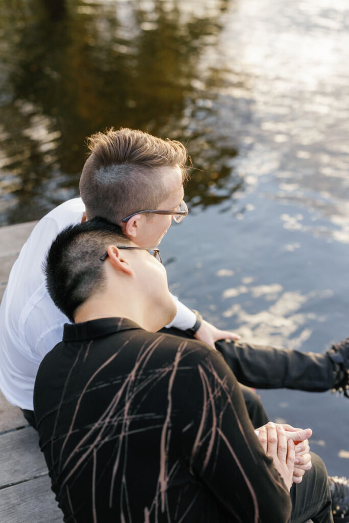 A person resting their head on their partner's shoulder as they sit on a small pier. 
