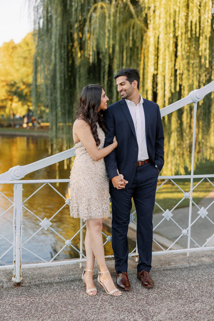 A couple holding hands and smiling at each other while standing in front of a lake. 