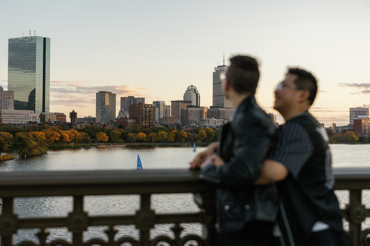 A couple slightly blurred as they are standing at a railing overlooking the water with a city skyline in the distance