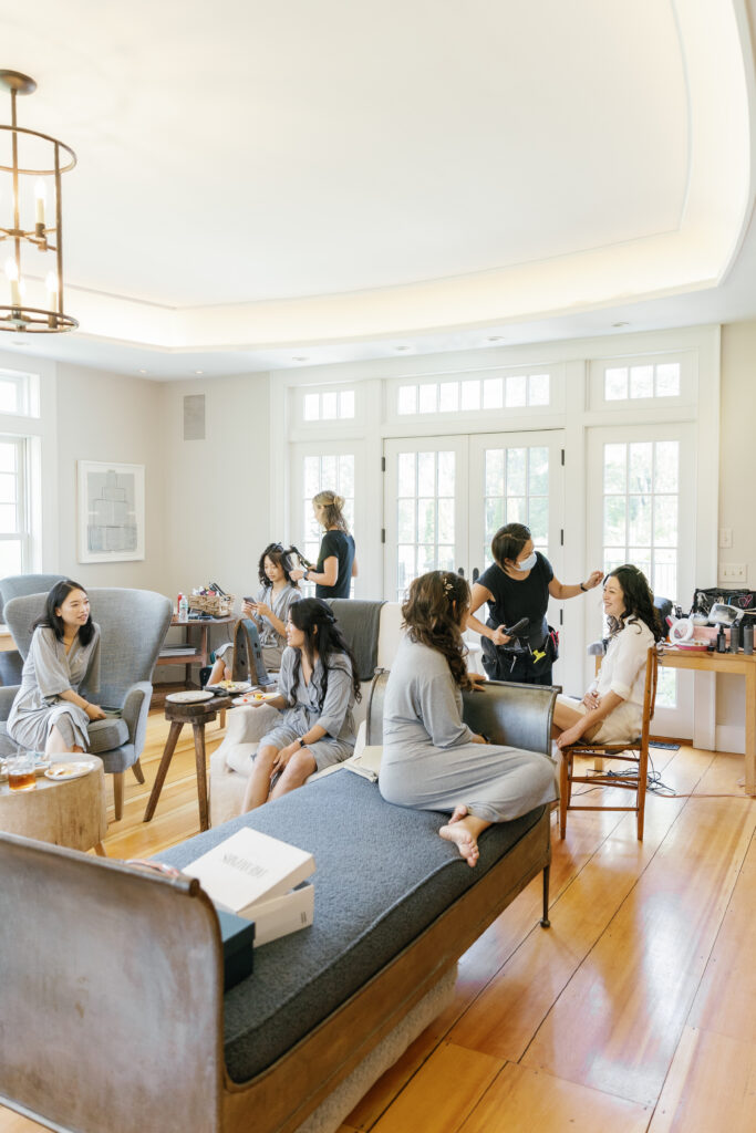 The bridal party chat and get their makeup done in a large, brightly lit room.